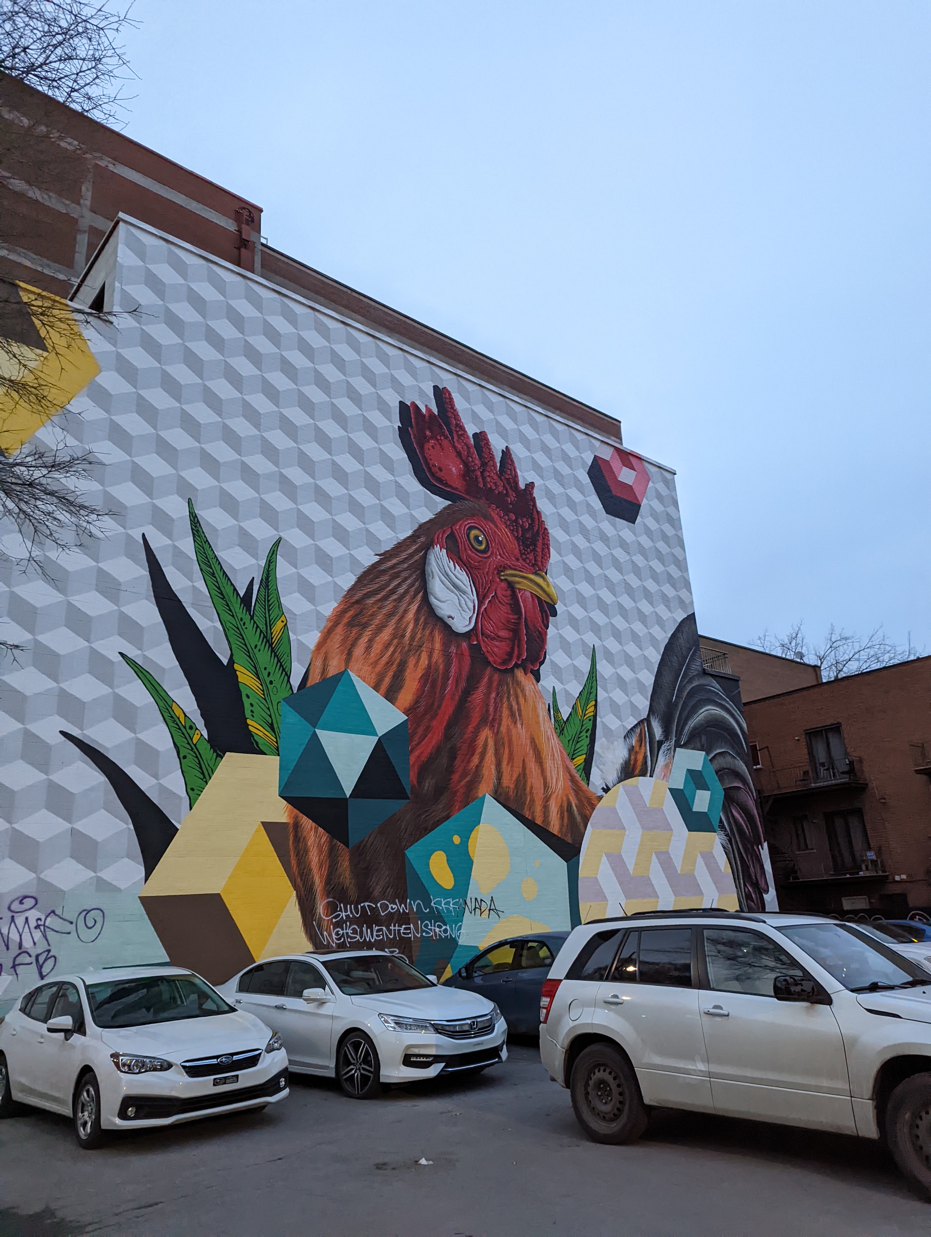 A mural of a chicken and some geometric shapes painted on the side of a two story brick building